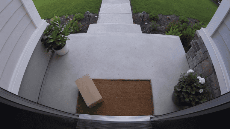 Front porch viewed from a doorbell camera perspective with wide-angle distortion, showing a package on the doormat in daylight.